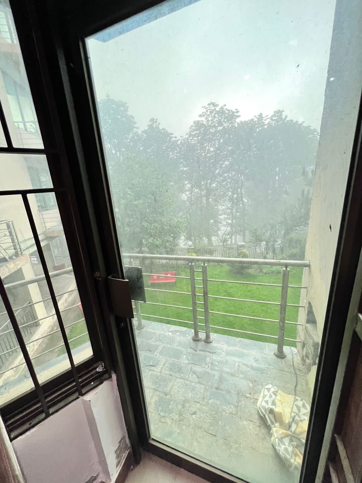 A glass door is shown, partially open, revealing a view of a garden with lush greenery and a few scattered trees. The ground is paved, and a cozy seating area can be seen to the side, creating a serene outdoor space.