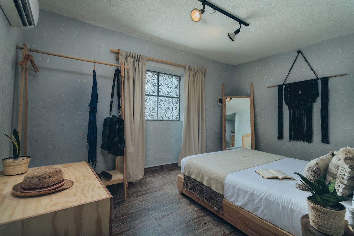 A serene sleeping area features a spacious bed covered with a light duvet. A minimalistic wooden dresser and a full-length mirror are positioned nearby. Natural light filters through a window adorned with sheer curtains, while plants add a touch of greenery to the space.