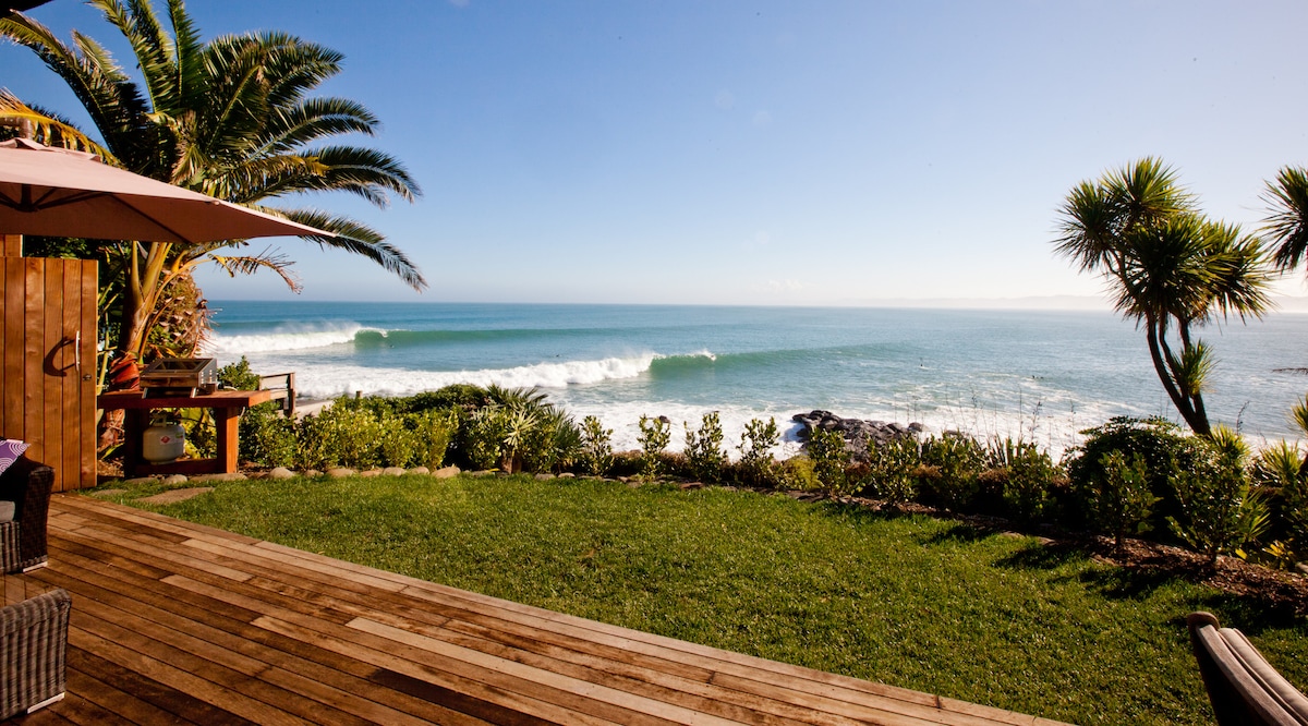A well-manicured lawn extends towards the ocean, showcasing the waves breaking gently on the shore. Tropical plants and palm trees frame the scene, with a wooden deck and outdoor furniture positioned to enjoy the coastal views.