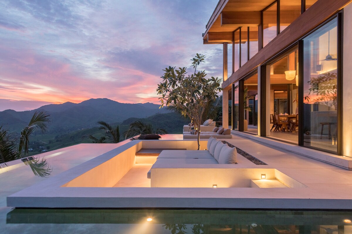 An expansive outdoor space features a serene infinity pool, seamlessly extending toward the horizon. Comfortable seating areas are arranged around the pool, with soft lighting accentuating the structure. The backdrop includes lush mountains under a vibrant sunset sky, highlighting the tranquil atmosphere.