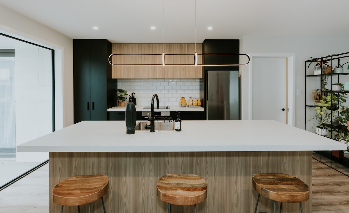 A modern kitchen features a spacious island with a light-colored countertop, paired with three wooden stools. Wooden cabinetry contrasts with black elements, and ample natural light enters through large sliding doors, enhancing the open layout.