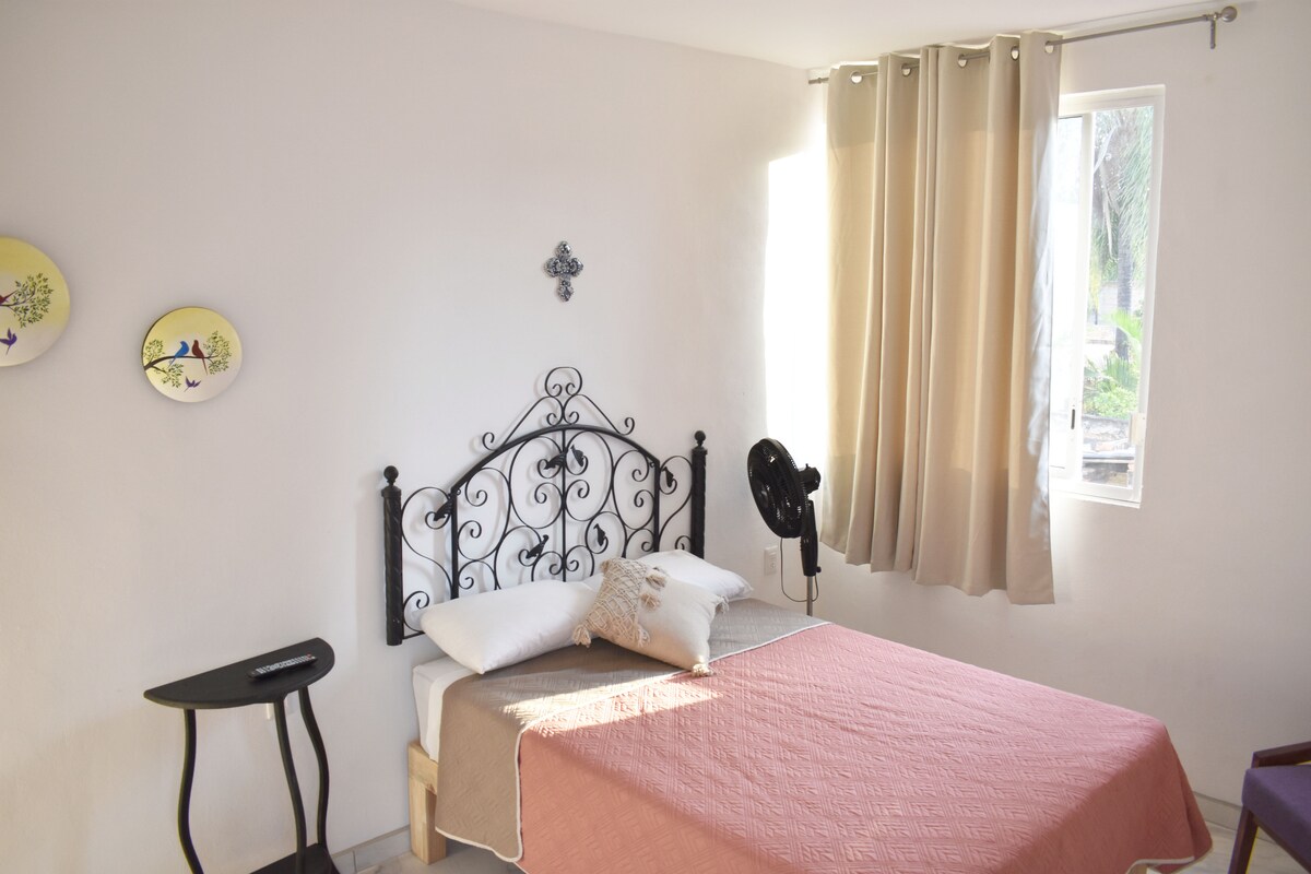 A cozy bedroom is presented with a decorative black metal bed frame and a light quilt draped across the bed. Natural light enters through a window adorned with soft curtains. A small, dark table stands beside the bed, complemented by framed wall art featuring floral designs.