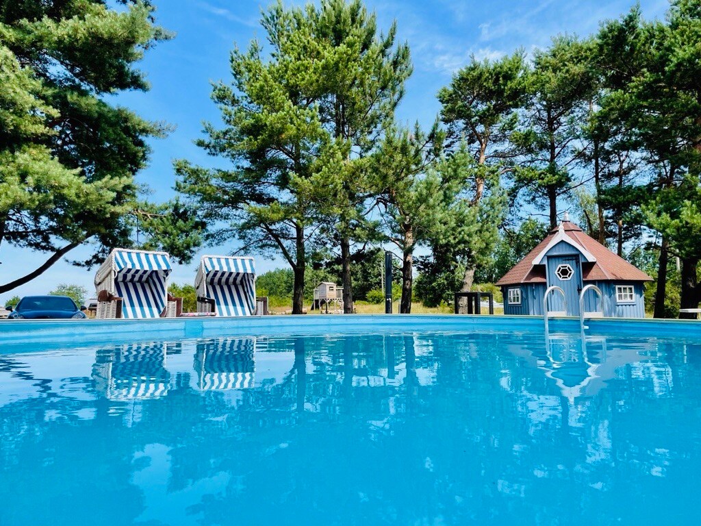 A sparkling outdoor pool reflects the clear blue sky, surrounded by lush green trees. Two striped lounge chairs are positioned near the water's edge, providing a serene relaxation space. A charming blue structure is situated in the background, adding character to the landscape.