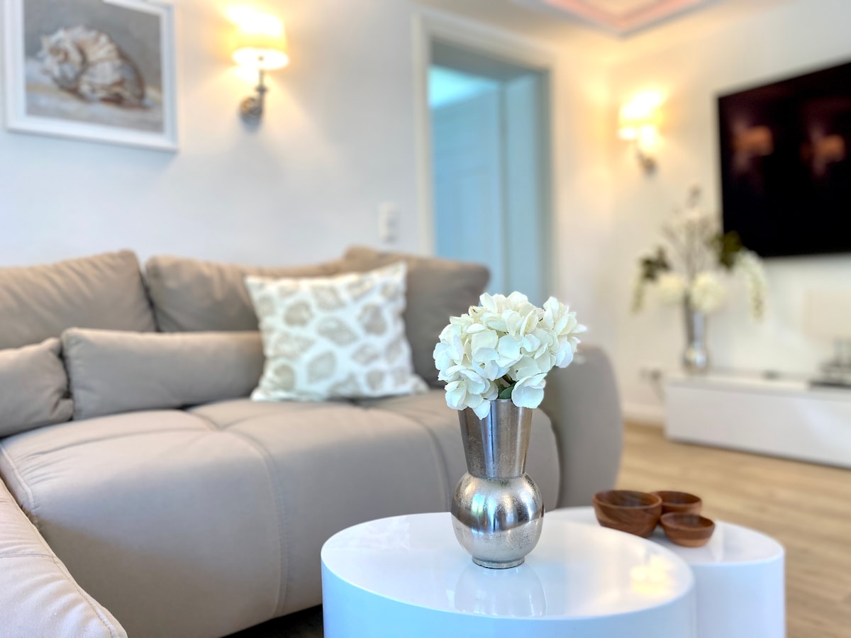 A soft-colored sofa is accompanied by a decorative pillow and a small white coffee table. A silver vase with white flowers is placed on the table alongside two wooden bowls. Soft lighting is provided by wall sconces, enhancing the serene environment.