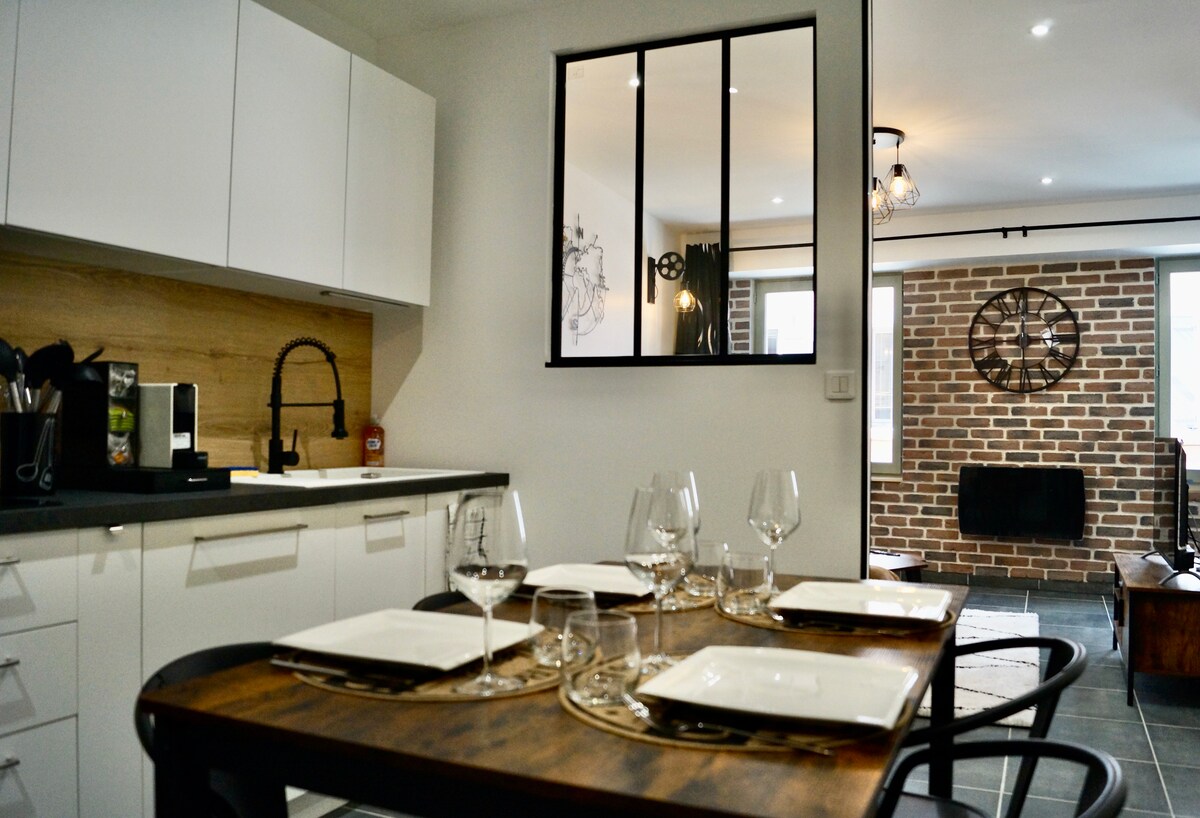 A modern kitchen is visible with white cabinetry and a wood backsplash. A dining table is set for four, featuring plates and glasses. A glass partition separates the kitchen from a cozy living area with exposed brick walls and decorative clock, enhancing the contemporary industrial style.