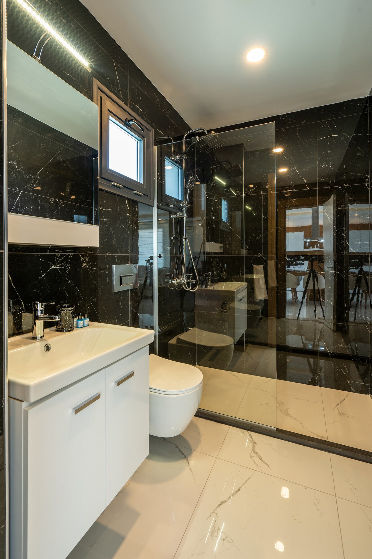 A modern bathroom is displayed, featuring sleek black marble walls and a large walk-in shower. A white vanity with storage is positioned below a mirror, while a minimalistic toilet is showcased. Natural light enters through a window, enhancing the contemporary design.