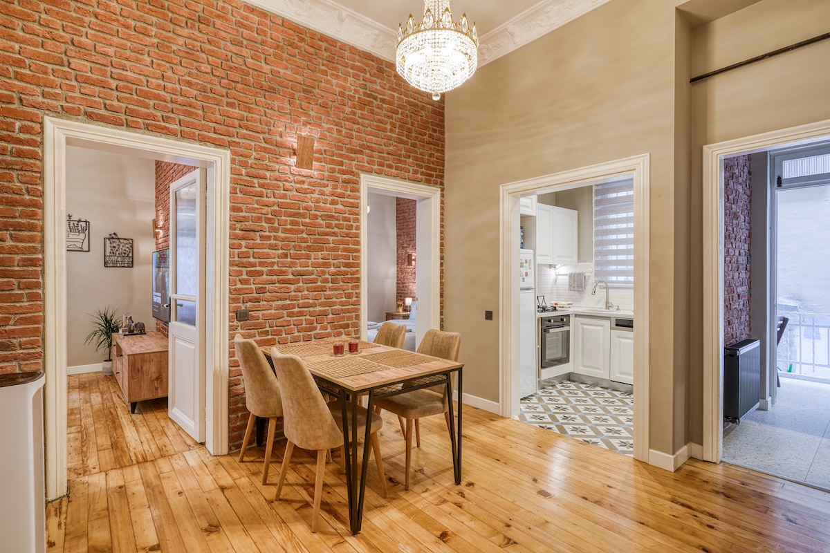A dining area features a wooden table surrounded by four upholstered chairs, set against a backdrop of exposed brick walls. Warm wooden flooring extends throughout the space. Light from a chandelier above adds a welcoming glow, while doorways lead to adjoining rooms.
