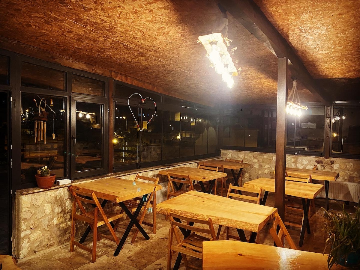 A spacious dining area features several wooden tables arranged with natural finishes. Large windows provide expansive views of the surroundings. Soft lighting from hanging fixtures creates a relaxed ambiance, inviting guests to enjoy their meals in comfort.