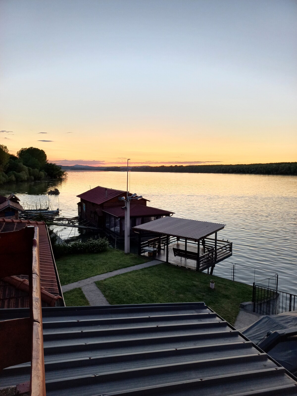 The image captures a tranquil river scene at sunset, with soft colors reflected on the water. A wooden structure with a roof extends over the water, alongside a grassy area. The view includes trees lining the riverbank, creating a serene backdrop.