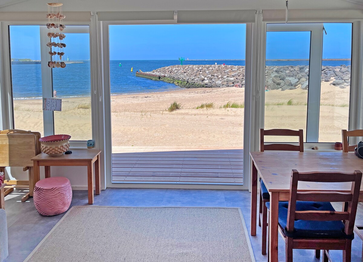 Large windows frame the tranquil seaside view, allowing natural light to fill the space. A dining table with wooden chairs is positioned nearby, while a cozy seating area is set against the wall. The sandy beach and calm water can be seen beyond the porch.