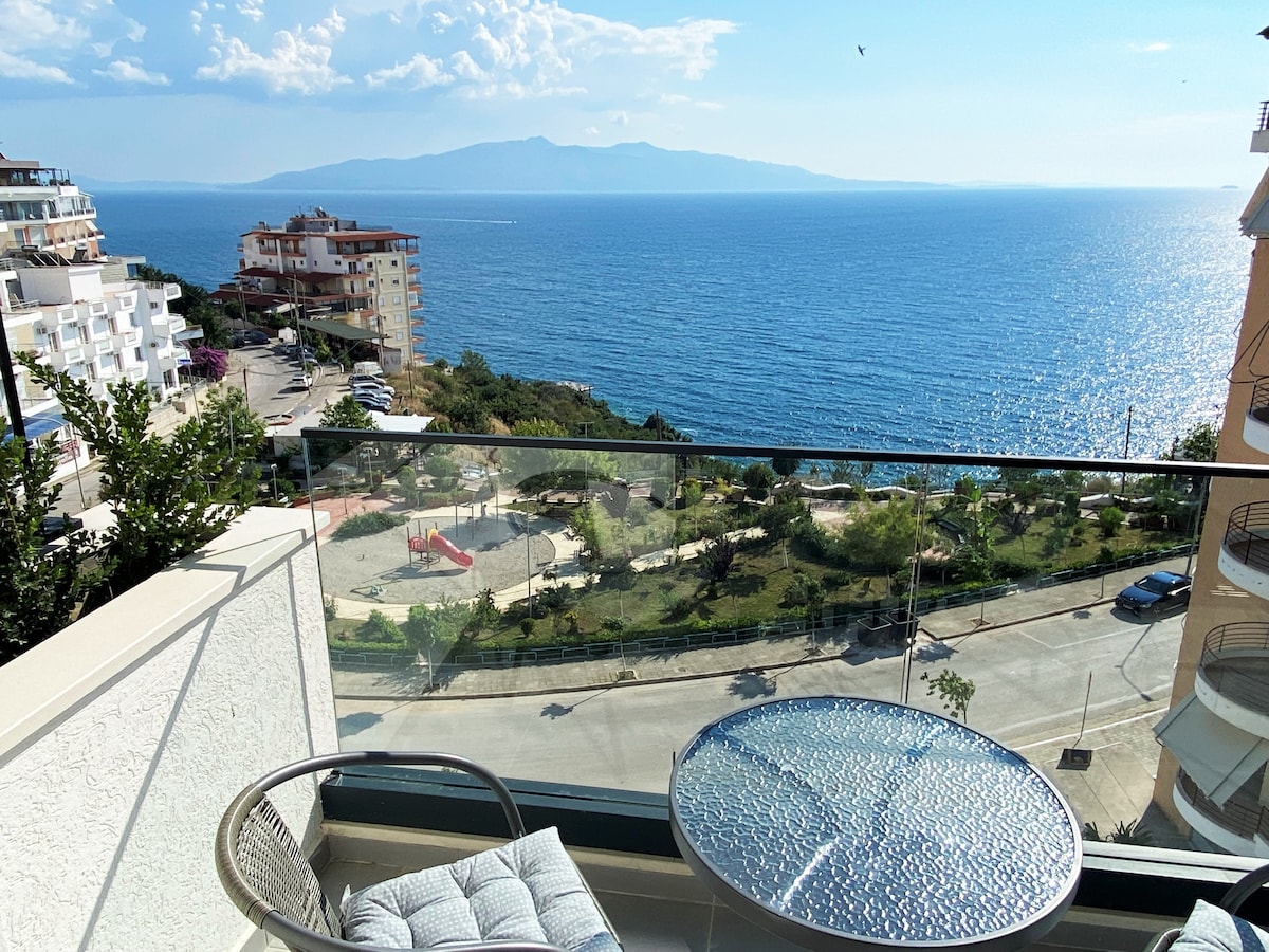 A scenic balcony view showcases a clear blue sea and distant mountains. A round glass table is paired with a comfortable chair, creating a relaxing outdoor space. Lush greenery and a playground are visible in the foreground, enhancing the tranquil coastal setting.