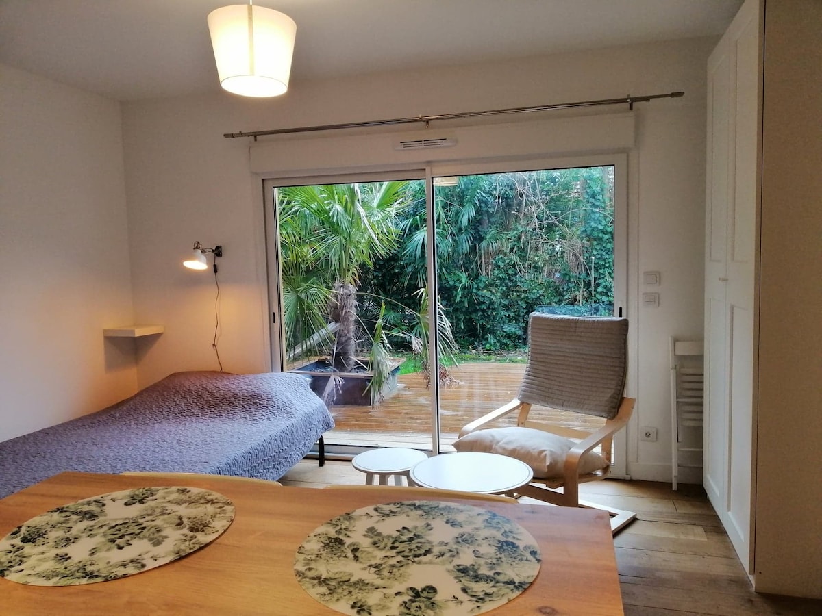 A bright studio space is highlighted by a spacious bed covered with a textured blanket. Natural light floods the room through large sliding glass doors, revealing a private terrace surrounded by greenery. A wooden table is set with decorative placemats, accompanied by a small table and a comfortable chair.