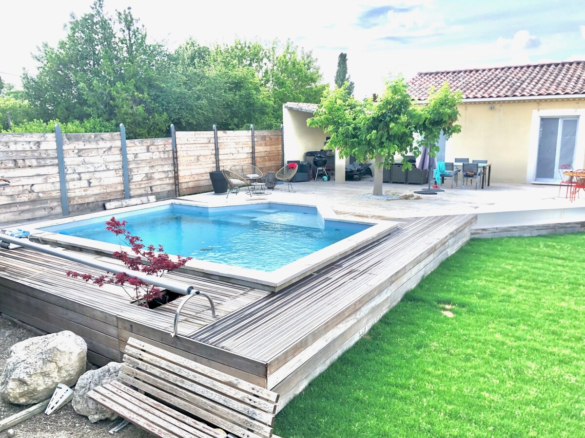 A backyard is revealed, featuring a rectangular salt-water pool surrounded by a wooden deck. Lush green grass complements the area, while a patio set and shaded seating are positioned nearby. Decorative plants and trees add touches of greenery to the enclosed space.