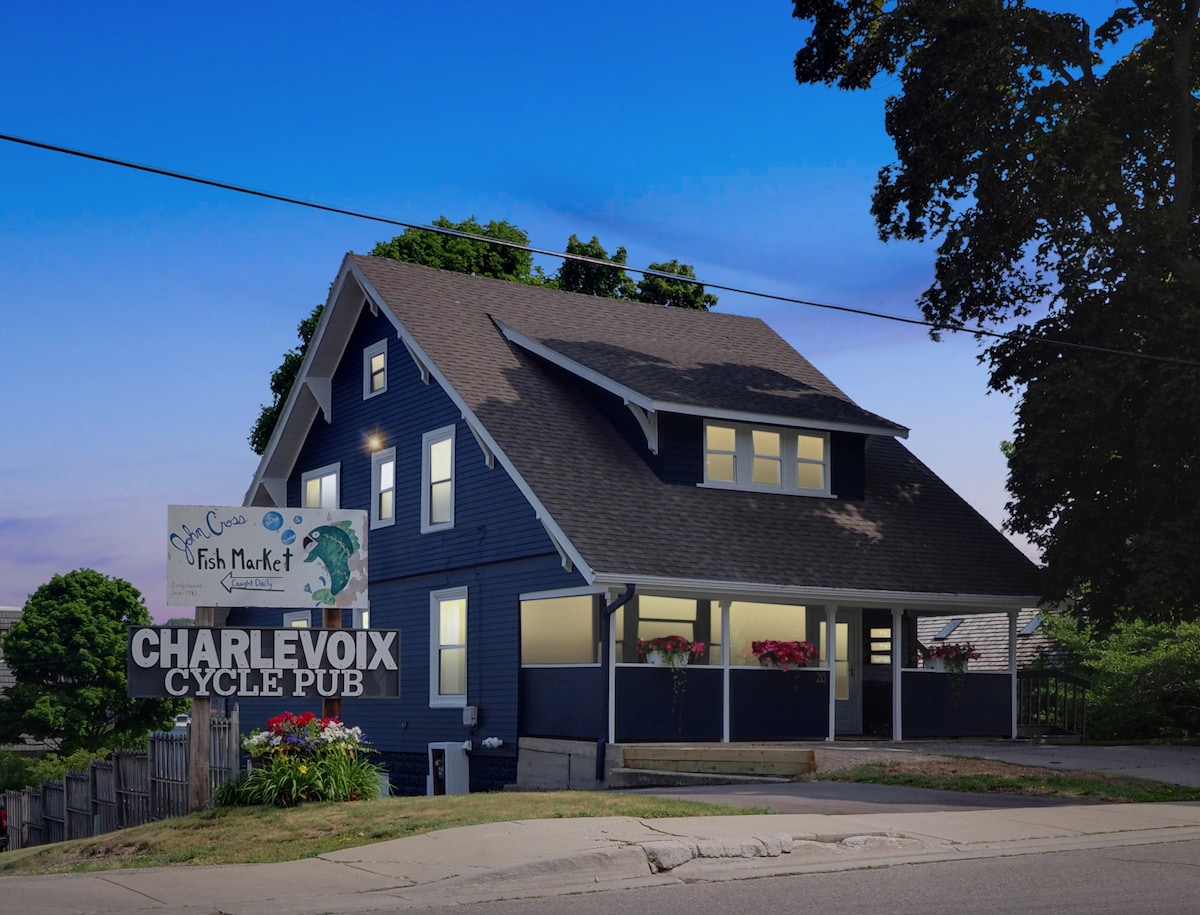 A charming blue house is seen from the street, featuring large windows and flower boxes. The front porch is welcoming, and a sign for Charlevoix Cycle Pub is prominently displayed along with an advertisement for a local fish market. Surrounding greenery adds to the inviting appearance.