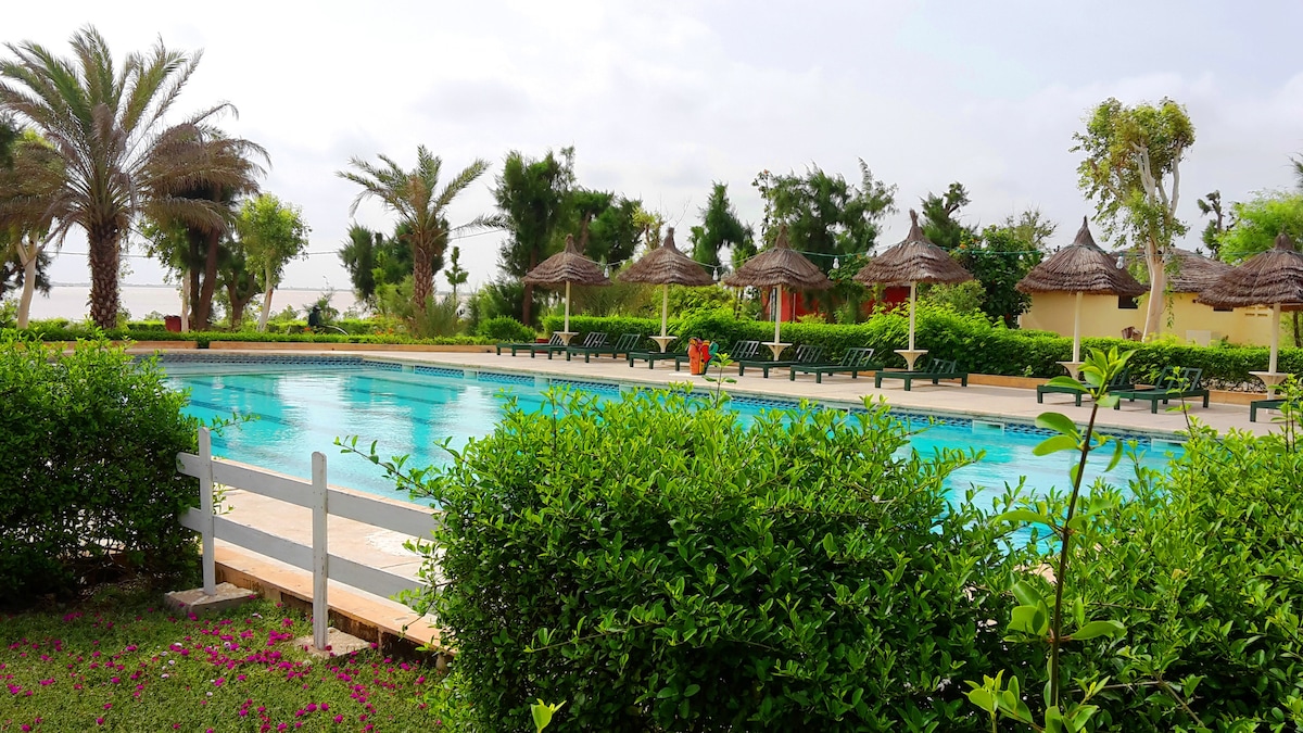 A large swimming pool is surrounded by neatly landscaped greenery, featuring palm trees and grass. Sun loungers are placed under thatched umbrellas along the poolside, offering shaded seating. The tranquil setting is ideal for relaxation.