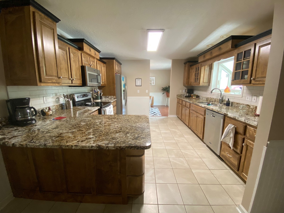 The kitchen features wooden cabinetry and granite countertops with ample prep space. Stainless steel appliances, including a refrigerator and microwave, are visible. A tile floor provides a neutral backdrop, and natural light enters through a nearby window, illuminating the open layout.