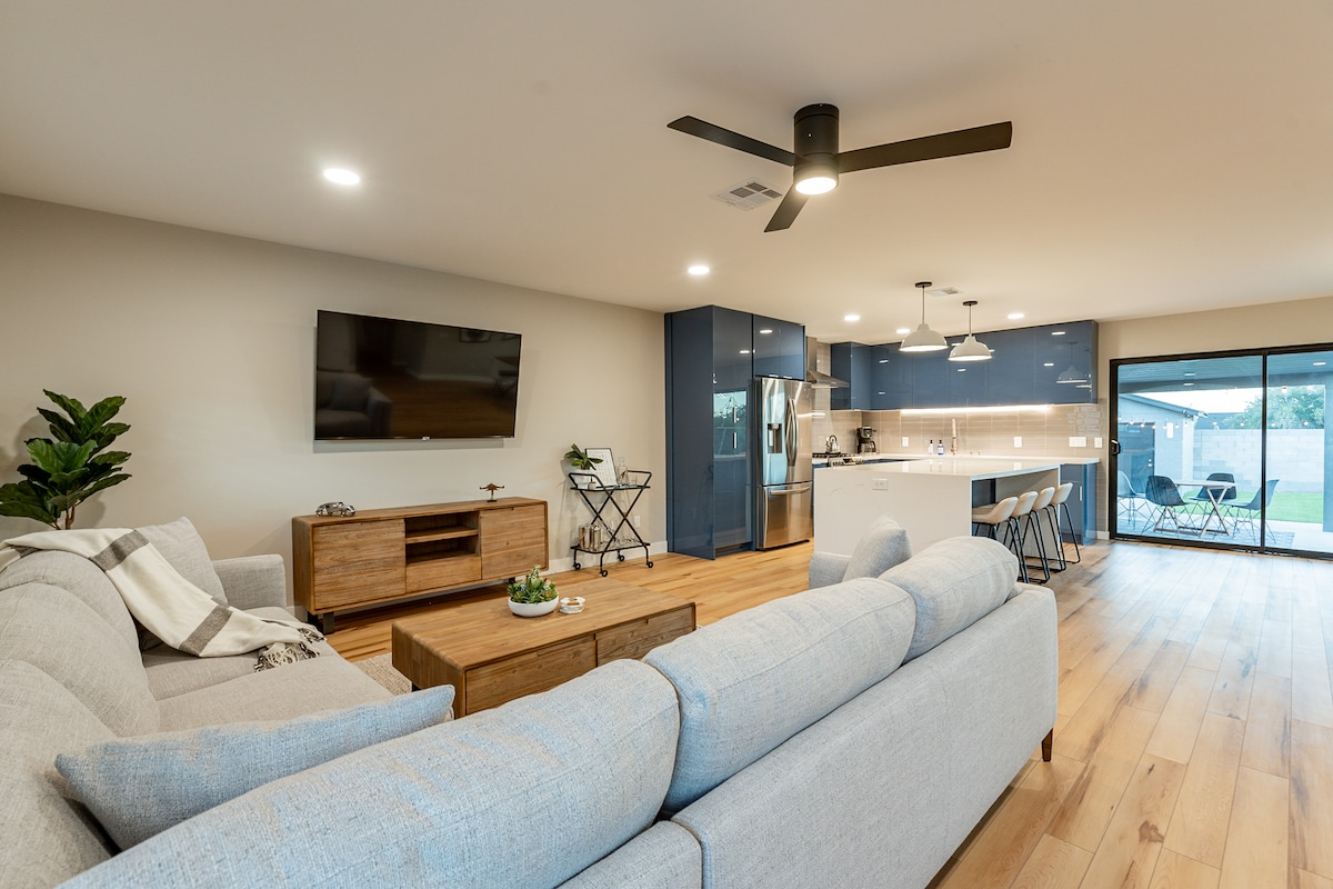 An open living area showcases a light-colored sofa facing a wall-mounted television. A wooden coffee table sits in front, accompanied by a decorative plant. The kitchen features modern blue cabinetry and a dining space with high-top chairs, leading to outdoor access through expansive glass doors.