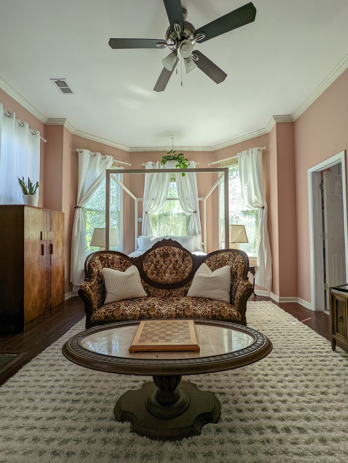 A bright sitting area features a patterned sofa with neutral-toned pillows, overlooking a sunlit window draped with light curtains. A round table sits in front, adorned with a chessboard. In the background, a spacious bed is visible through the open doorway.
