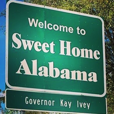 A green road sign features the text 'Welcome to Sweet Home Alabama' prominently displayed in white letters. Below it, the name 'Governor Kay Ivey' is shown, indicating the sign's official status. Surrounding foliage is visible, creating a natural backdrop.