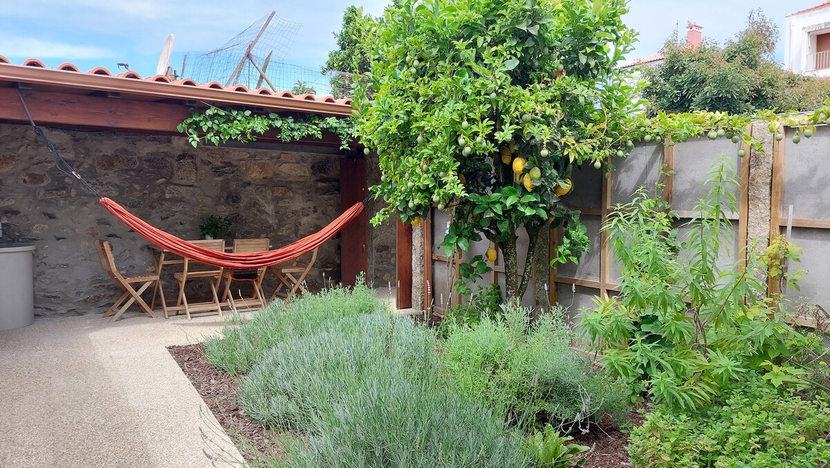An outdoor space features a hammock suspended between wooden beams, a small wooden table surrounded by chairs, and lush greenery including lemon trees and herb plants. Stone walls provide a rustic backdrop, while a terracotta roof adds to the charm of the area.