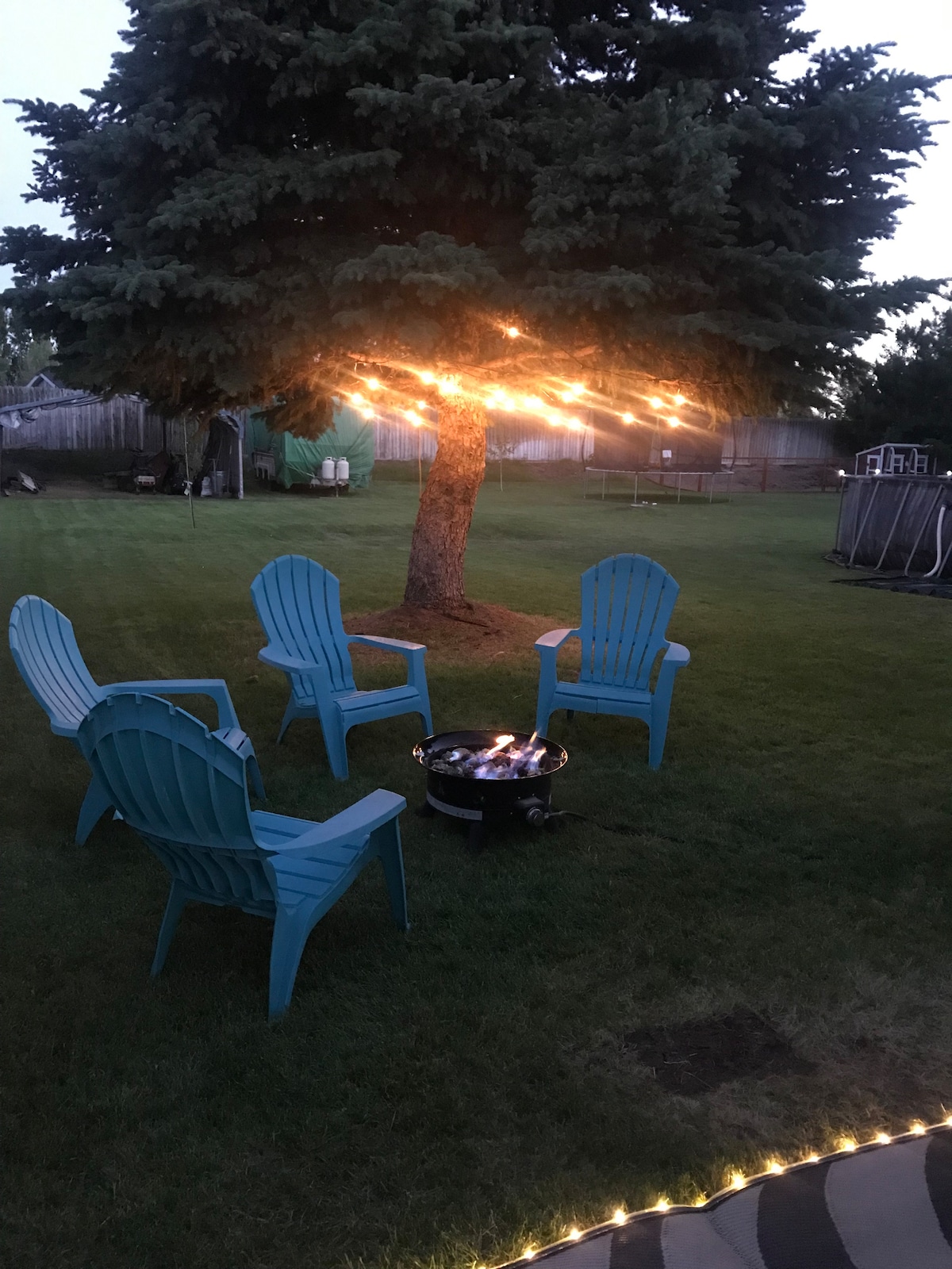 A serene outdoor area features a cozy gas fire pit surrounded by four blue Adirondack chairs. Soft lighting is strung from a tree above, providing a warm glow in the grassy setting. The space is ideal for relaxation and gathering under the evening sky.