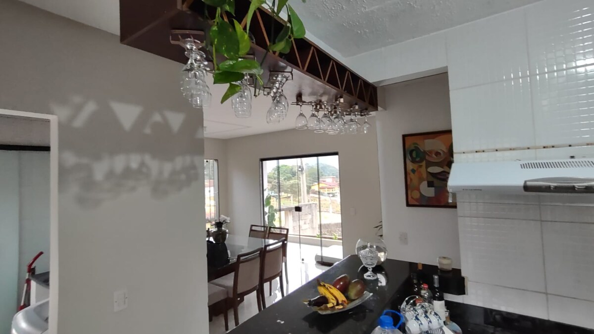 A modern kitchen features a bar with decorative glassware suspended above. Natural light fills the space through large windows overlooking a distant view. A dining table with wooden chairs is visible, complementing a minimalist decor style.