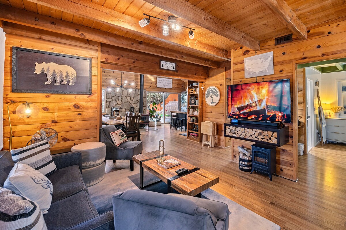 The open living area features warm wooden walls and a cozy seating arrangement, including a sectional couch and wooden coffee table. A large television displays a fake fireplace, and dining spaces are visible in the background, inviting guests to gather together.
