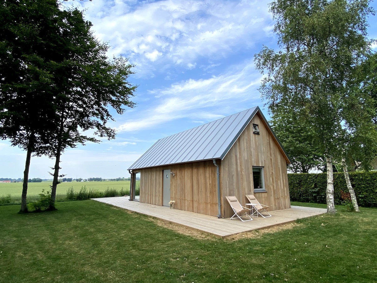 A contemporary wooden summerhouse is shown with a sloped metal roof, situated on a grass lawn. Two lounge chairs are positioned on a wooden deck in front, surrounded by trees and expansive views of the fields in the background under a partly cloudy sky.