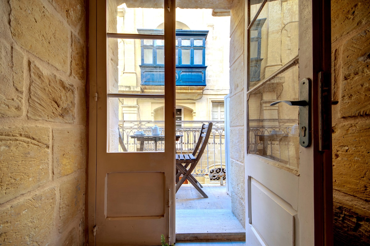 A view from the entrance showcases a private balcony with wooden furniture. Blue shutters decorate the balcony, adding a touch of color against the stone walls. Natural light flows through the glass doors, highlighting the traditional architecture of the maisonette.