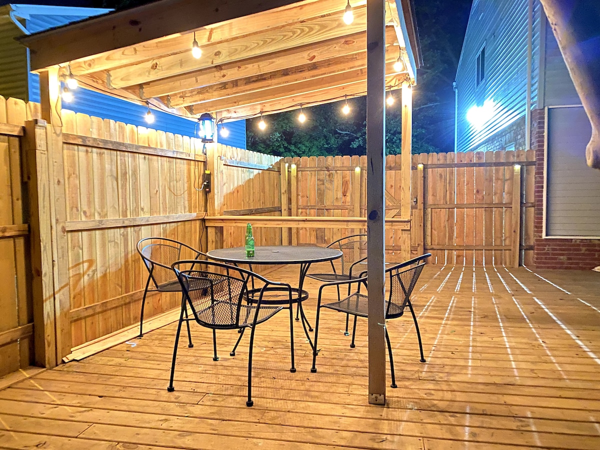 A fenced backyard features a wooden deck with string lights overhead. A round table is surrounded by four black wire chairs, creating a cozy seating area for outdoor gatherings. The warm glow from the lights adds a welcoming touch to the space.