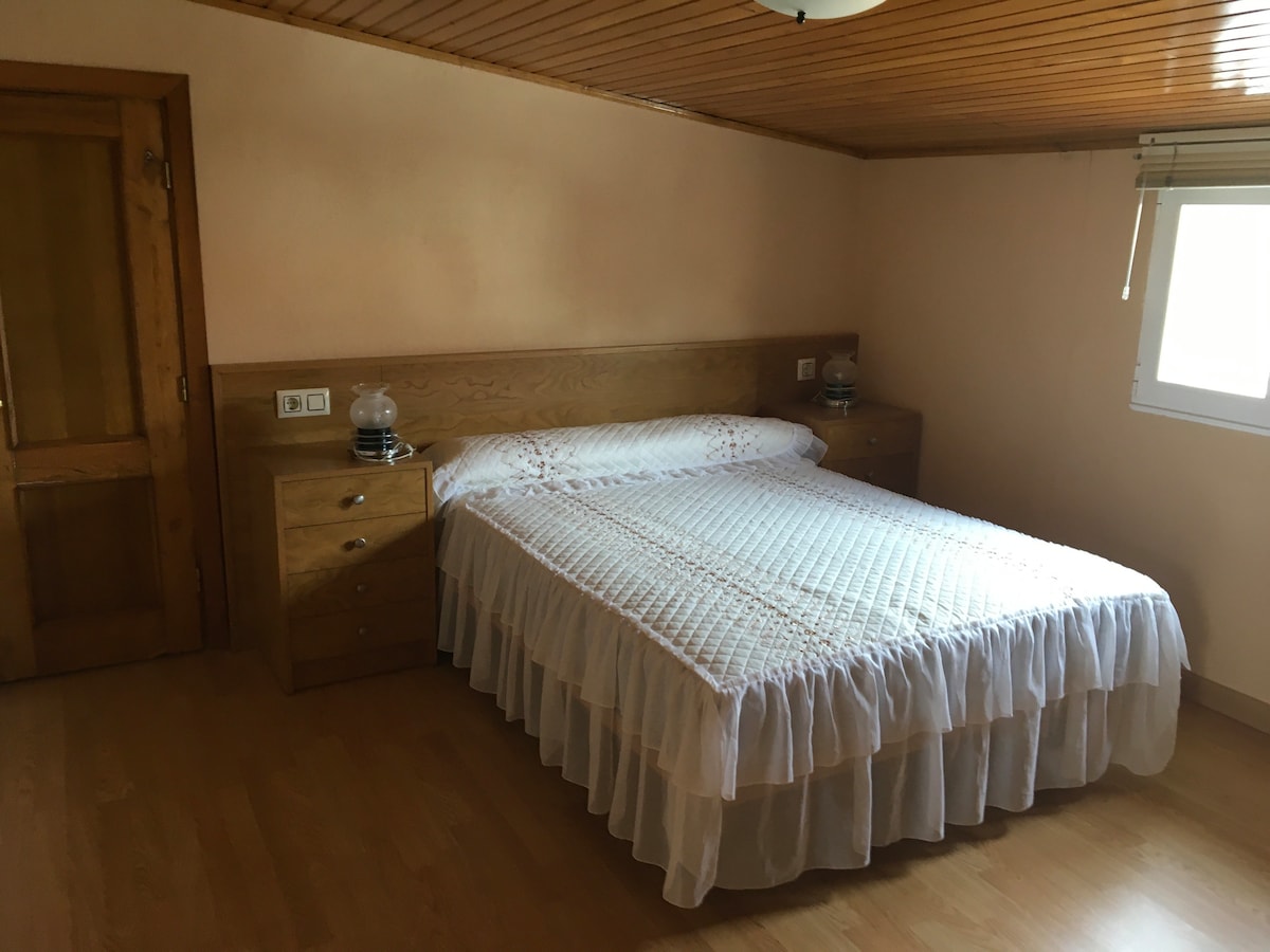 A spacious bedroom features a large bed with a white bedspread and a decorative ruffle. Wooden nightstands are positioned on either side, each with a lamp. A small window allows natural light to enter, and the ceiling showcases wooden paneling.