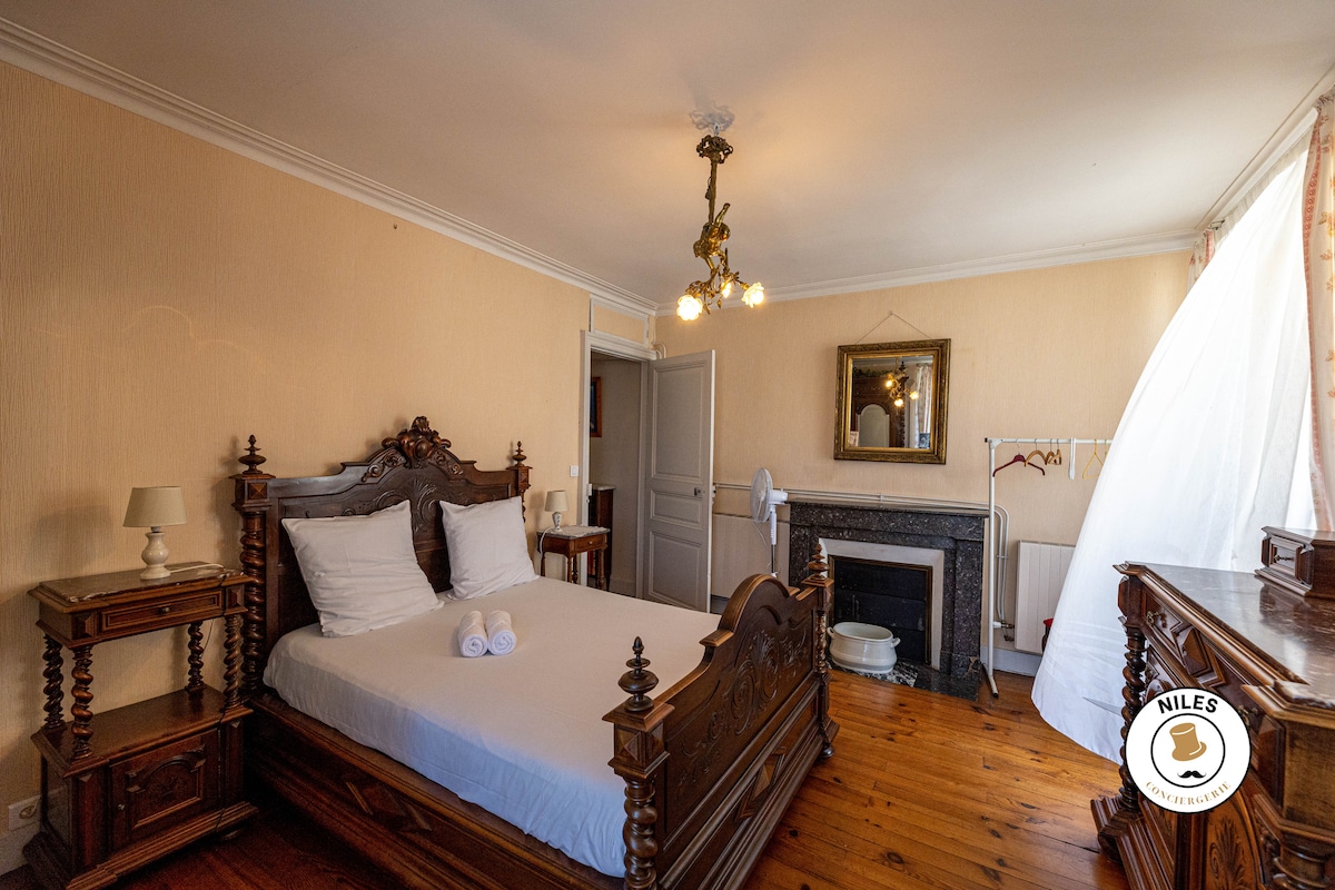 The room features a classic wooden bed with a white bedding set and neatly arranged towels. A vintage-style chandelier hangs from the ceiling, providing soft illumination. An ornate mirror and a decorative fireplace enhance the traditional decor, complemented by wooden flooring.