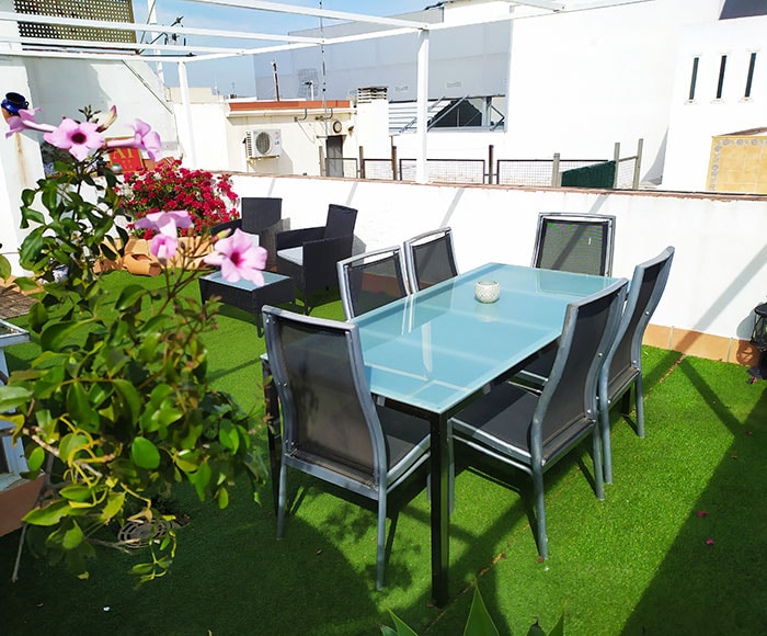 An outdoor seating area features a glass-top dining table surrounded by six black chairs. Lush green grass covers the ground, while pink flowers are visible in the foreground, adding color to the inviting space. A relaxed environment for outdoor dining is presented.