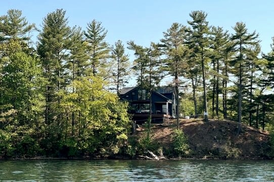 A modern cabin sits elevated above the water, surrounded by tall trees and lush greenery. The large deck is visible, providing outdoor space from which to enjoy the scenic views. The calm lake reflects the natural surroundings, enhancing the serene atmosphere.
