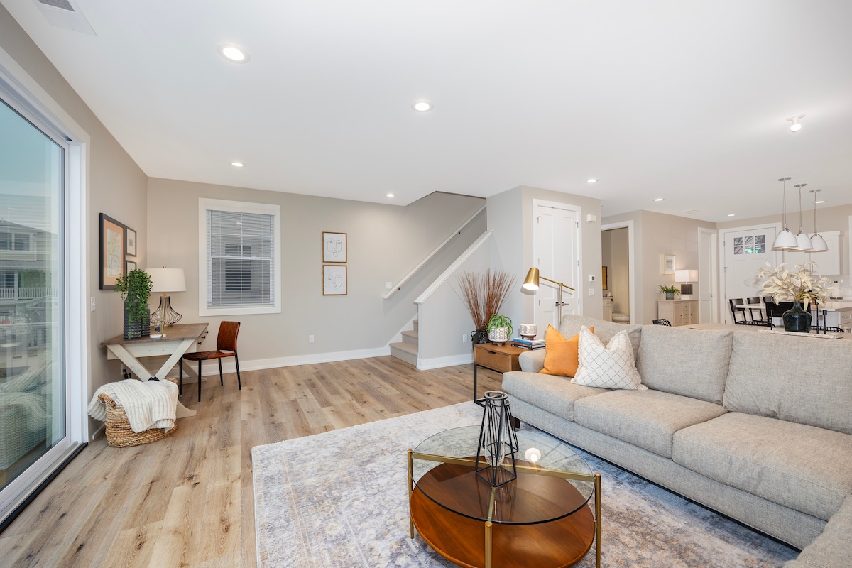 An open living space features a spacious sectional sofa paired with a round coffee table. A desk with a chair is positioned beside a large window. Light hardwood flooring extends throughout, complemented by a decorative area rug. Neutral tones are used on the walls, enhancing the airy feel.