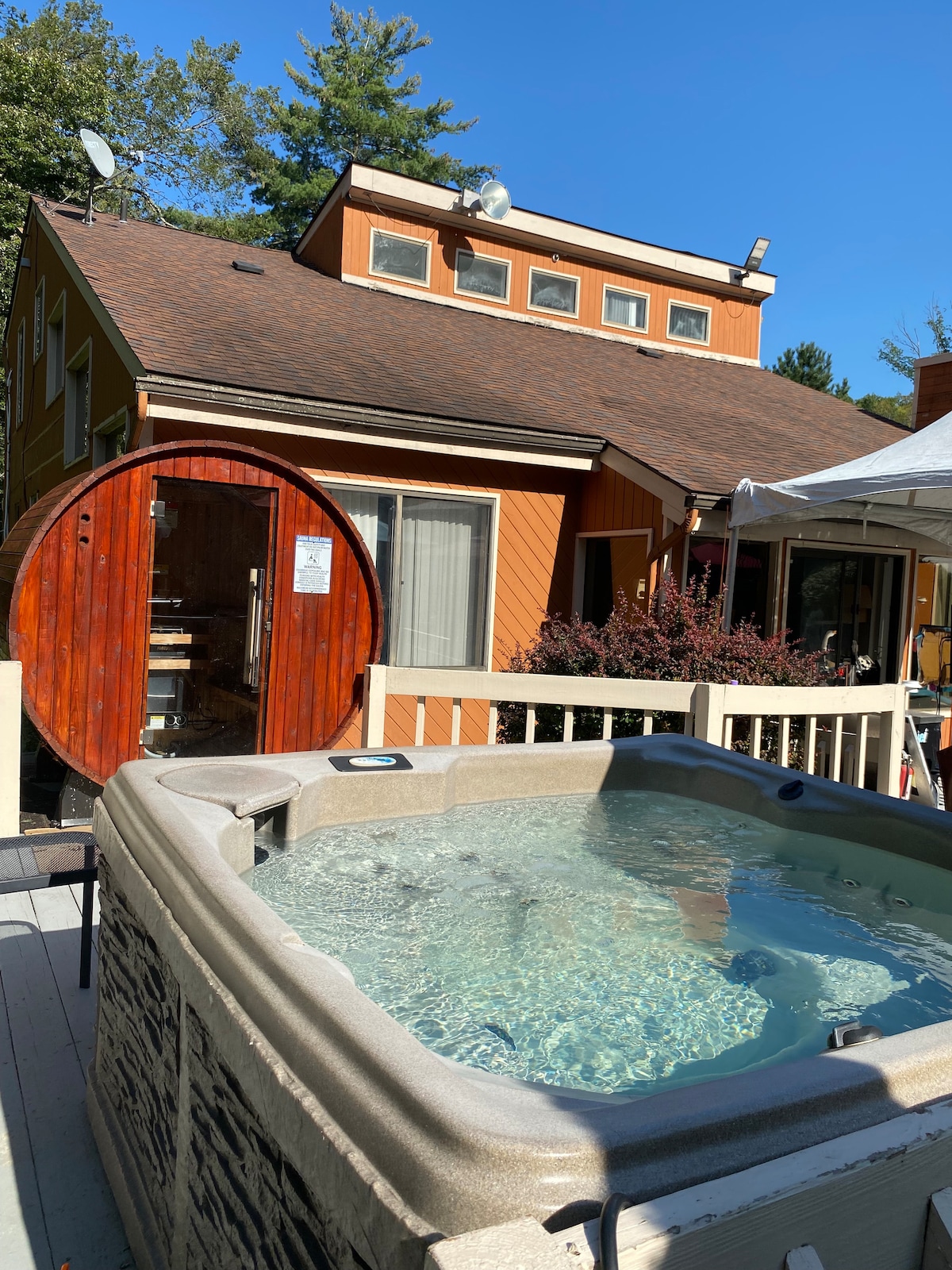 A spacious hot tub is positioned on a wooden deck, glistening under sunlight. In the background, a charming building features warm orange siding and large windows, alongside a wood-fired sauna adorned with a round entrance.