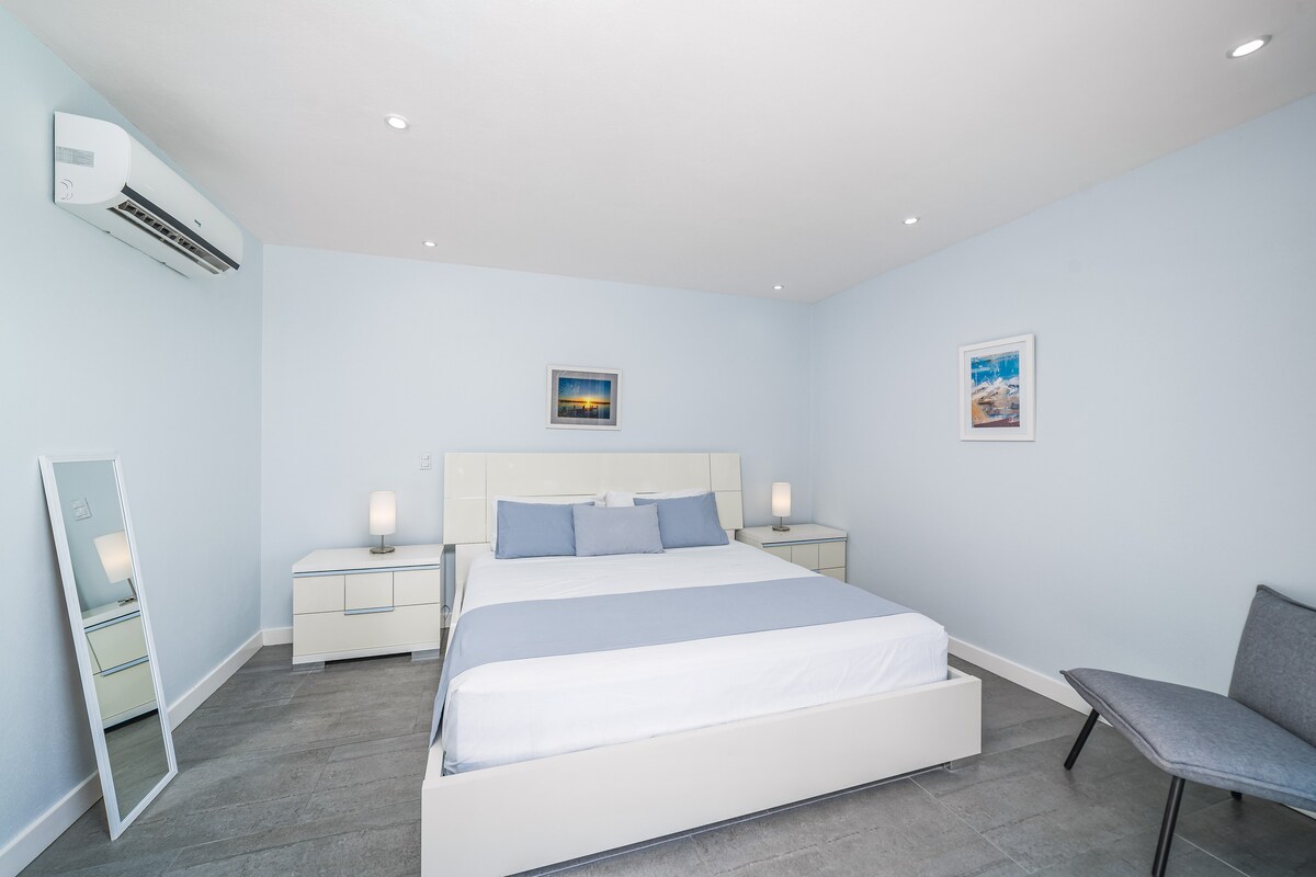 ห้องนอนใหญ่ที่เงียบสงบนี้ให้บรรยากาศที่สงบด้วยเฟอร์นิเจอร์สีขาวและโทนสีที่ผ่อนคลาย หน้าต่างบานใหญ่ช่วยให้แสงธรรมชาติส่องเข้ามาในที่พักและเข้าถึงสระว่ายน้ำได้โดยตรงและวิวสวนที่สวยงาม