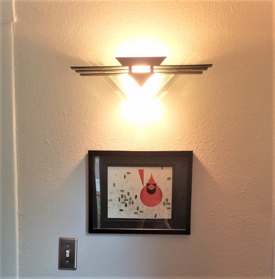 A wall-mounted light fixture illuminates the area, casting a warm glow. Below, a framed artwork featuring a stylized red cardinal is positioned against a light-colored wall. The artwork is surrounded by decorative elements, contributing to the overall aesthetic of the space.