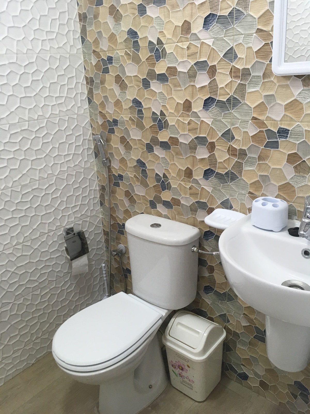 A clean bathroom features a modern toilet and a compact sink, accompanied by a decorative wall adorned with textured tiles in warm tones. A small waste bin and a shower fixture are also visible, providing essential amenities in this well-maintained space.