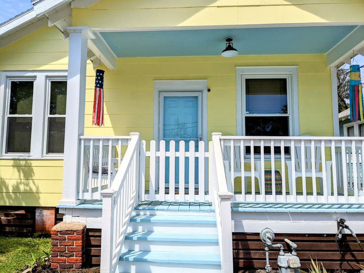 The exterior of a vintage coastal cottage is presented, featuring a light yellow façade. A white picket fence borders the front porch, which includes two white chairs and a welcome blue door. Decorative items, including an American flag and colorful wind streamers, enhance the cheerful atmosphere.