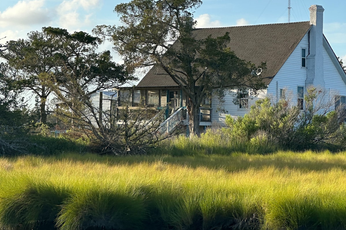 A cozy cottage built in 1848 is set amidst lush green grass in the foreground. Large trees frame the home, which features a spacious deck and a sloped roof. The tranquil setting captures the charm of rustic living near the water.