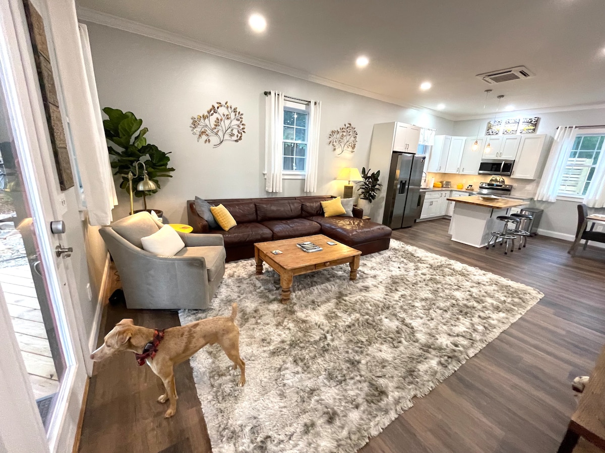 A cozy living area is highlighted by a leather sectional sofa, accompanied by a light armchair. Natural light fills the room, which features a plush rug and wooden coffee table. A dog stands near the door, while the fully equipped kitchen is visible in the background.