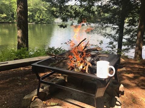 A campfire burns brightly within a black fire pit, surrounded by a stone circle. A white coffee mug rests on the edge of the pit. The serene lake is visible in the background, framed by lush trees and a clear sky.