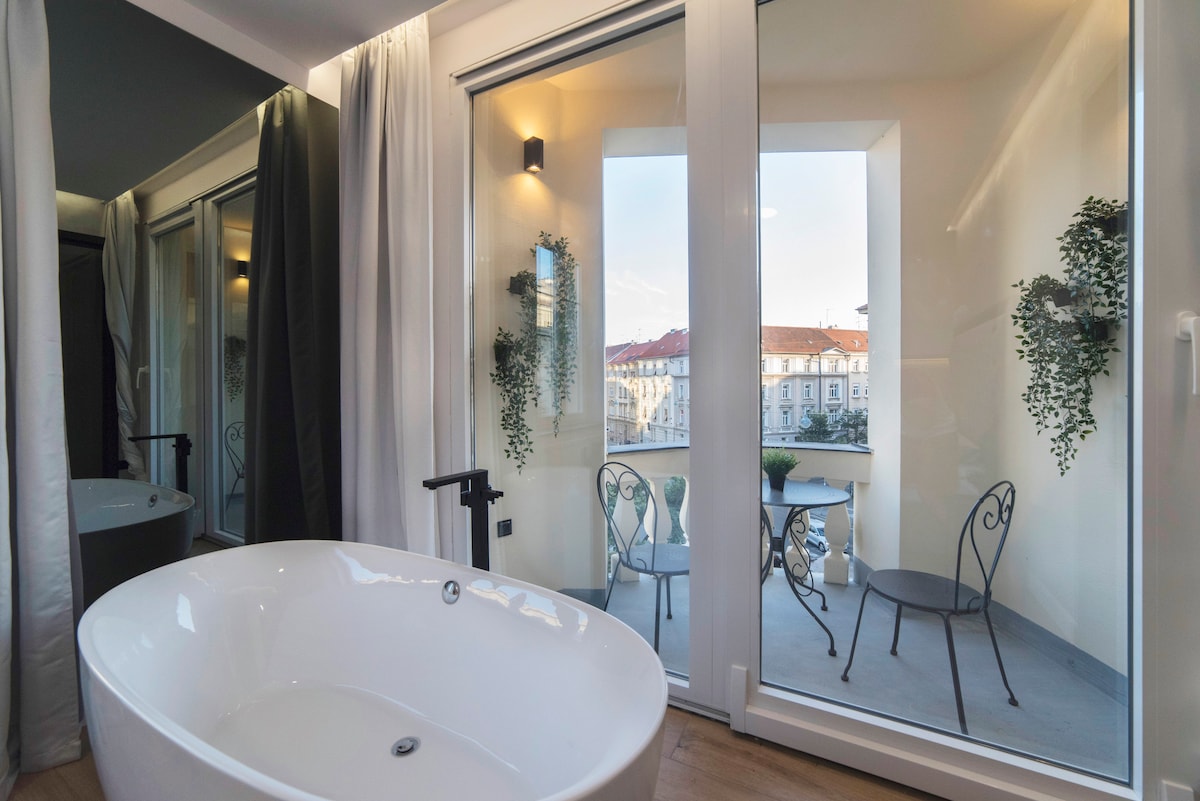 A freestanding bathtub is positioned near large glass doors, which lead to a romantic balcony. The balcony features two wrought iron chairs and potted plants, offering a view of the surrounding urban environment.