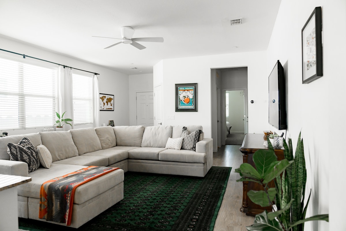 A spacious living room is highlighted by a large sectional sofa, complemented by a decorative throw and patterned cushions. Natural light fills the room through multiple windows, and a flat-screen TV is mounted on the wall. A rug covers the floor, enhancing the cozy atmosphere.
