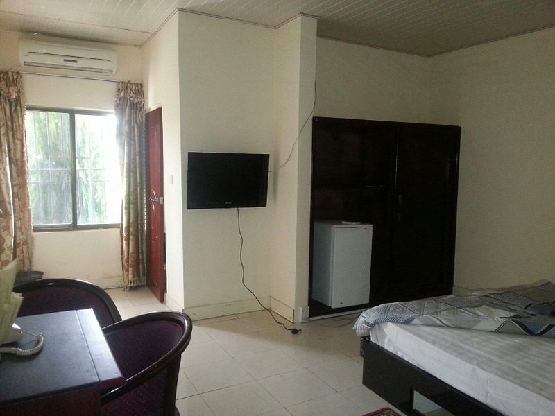 A spacious room features a desk with two chairs, a flat-screen television mounted on the wall, and a mini-fridge. Soft sunlight enters through a large window beside beige curtains. A bed is positioned in the corner, with light-colored bedding contrasting the dark furniture.