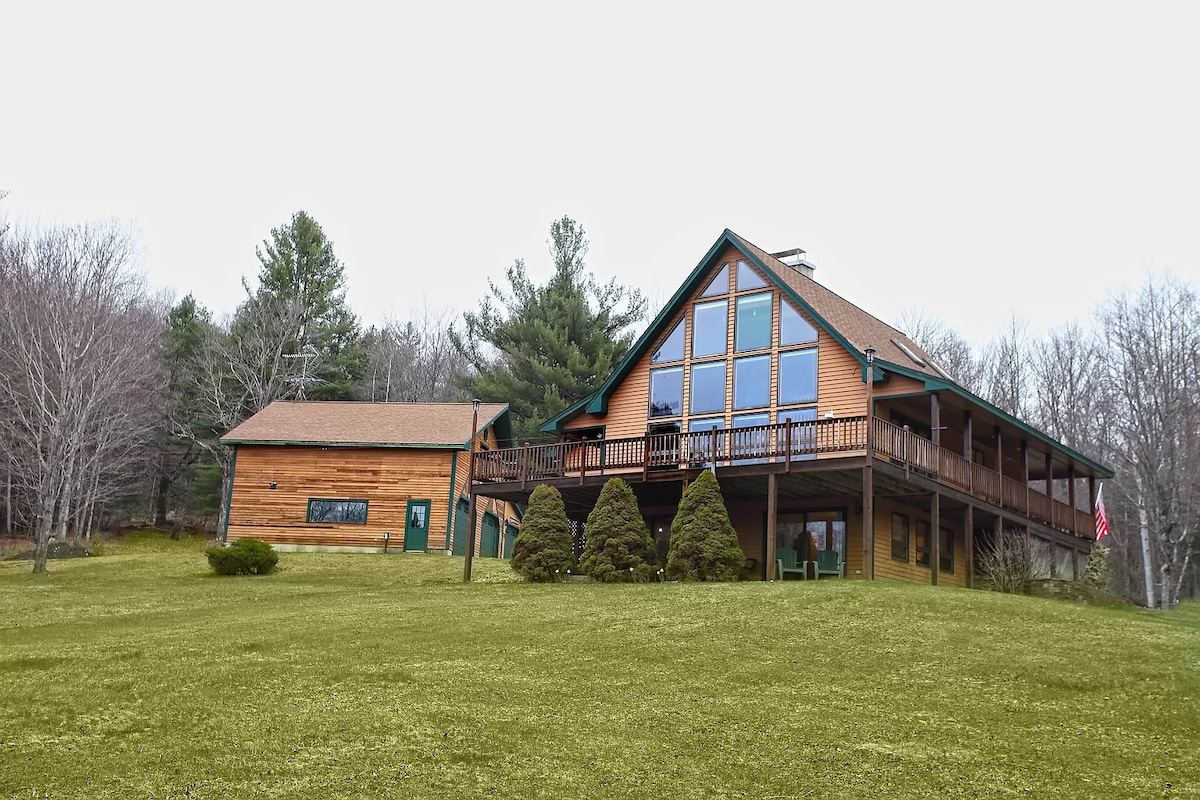 A chalet-style home is surrounded by lush greenery, featuring a large three-sided wrap-around deck. Tall windows dominate the front, allowing ample natural light. To the side, a wooden garage complements the main structure, set against a backdrop of trees and an expansive lawn.