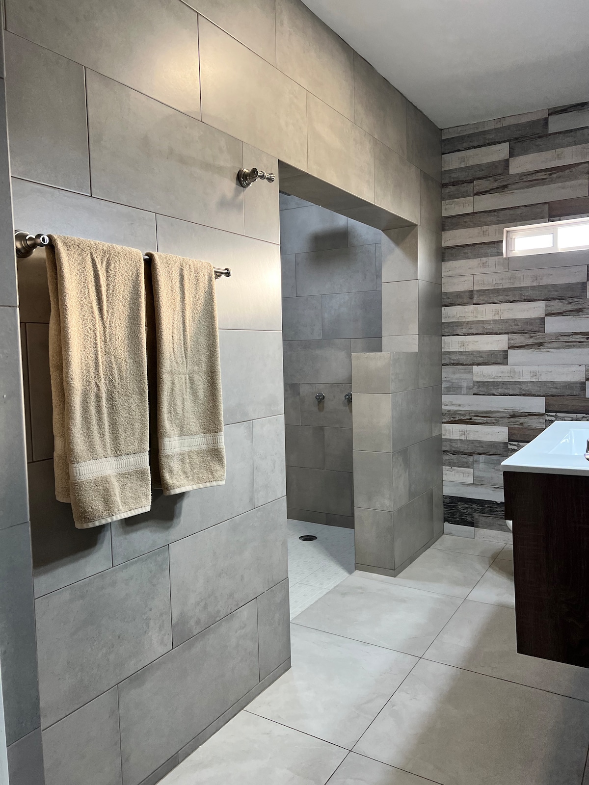 A modern bathroom features large gray tiles on the walls and floor, with a separate shower area visible beyond an archway. Two neatly hung towels are placed on chrome hooks. A sleek vanity with a white sink is positioned adjacent to the shower.