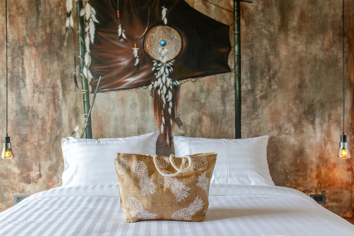 A spacious bed is positioned against a textured wall adorned with a large mural featuring natural elements. Two decorative pillows are arranged at the head of the bed, accompanied by a woven bag placed thoughtfully on the covers. Suspended bulbs add subtle illumination.