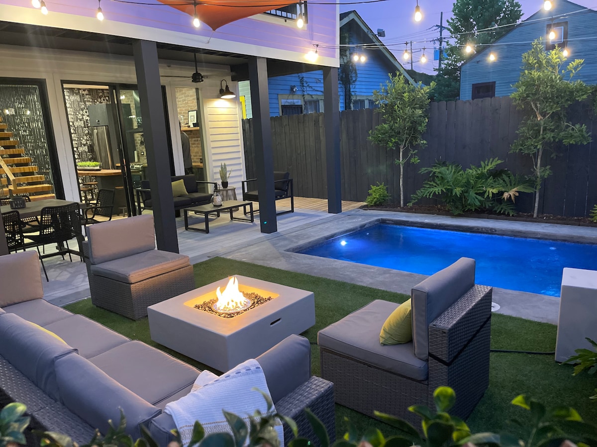 An outdoor entertainment area features comfortable seating around a fire pit table, with a serene pool nearby. The space is illuminated by string lights and includes lush greenery, offering a cozy atmosphere for relaxation and gatherings.